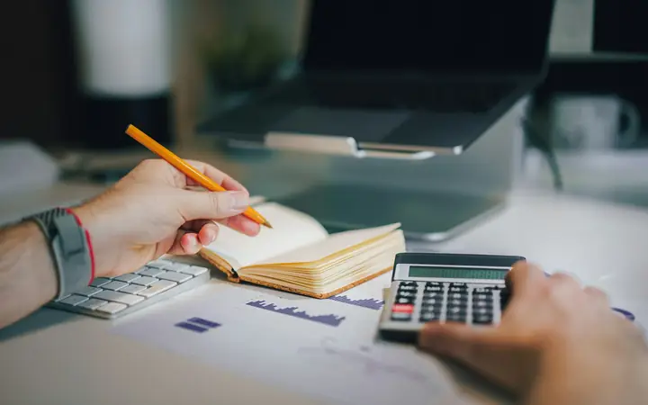 A person working out a budget with a calculator, a notebook and a laptop
