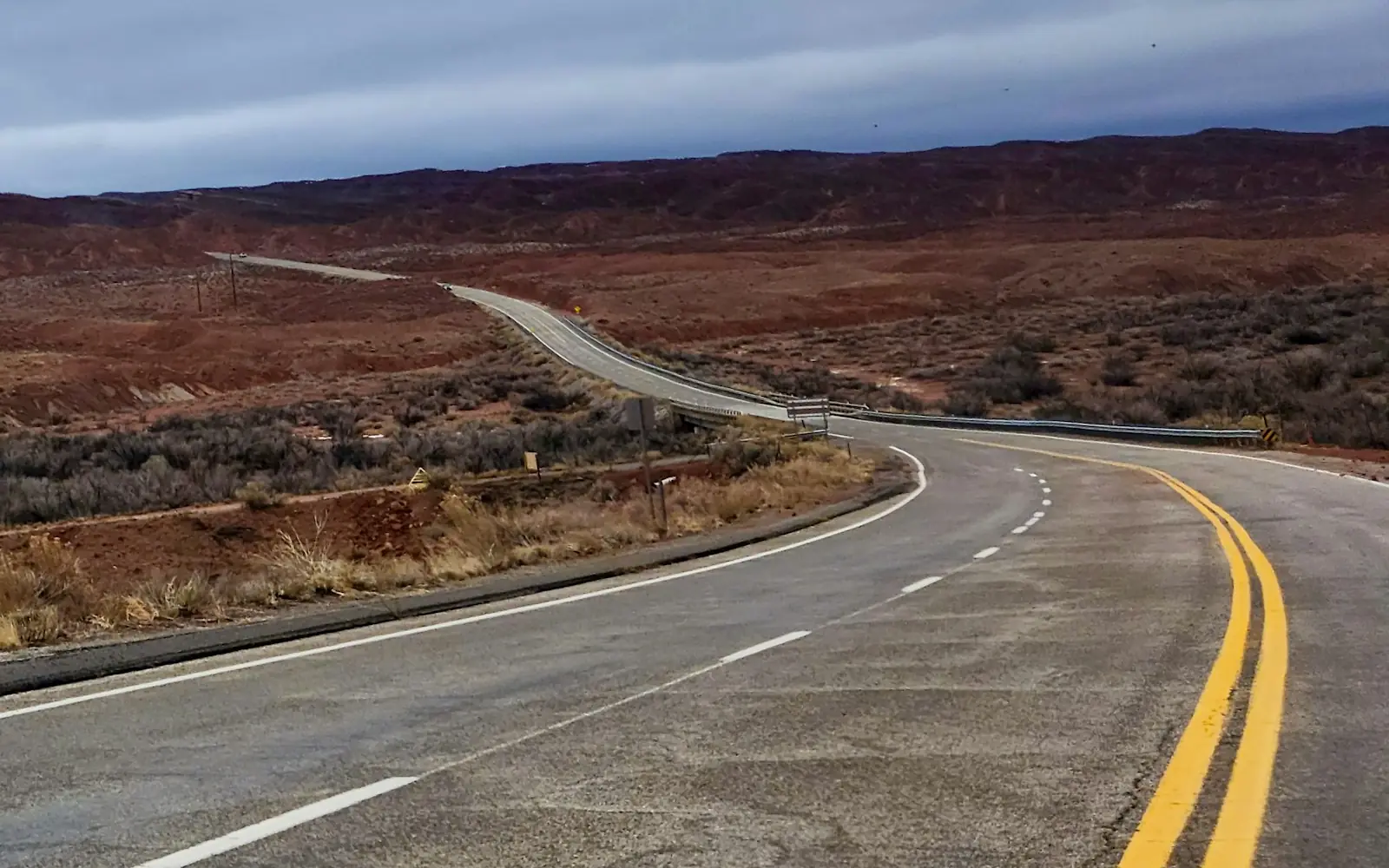 A two-lane road winding through red desert hills under a cloudy sky